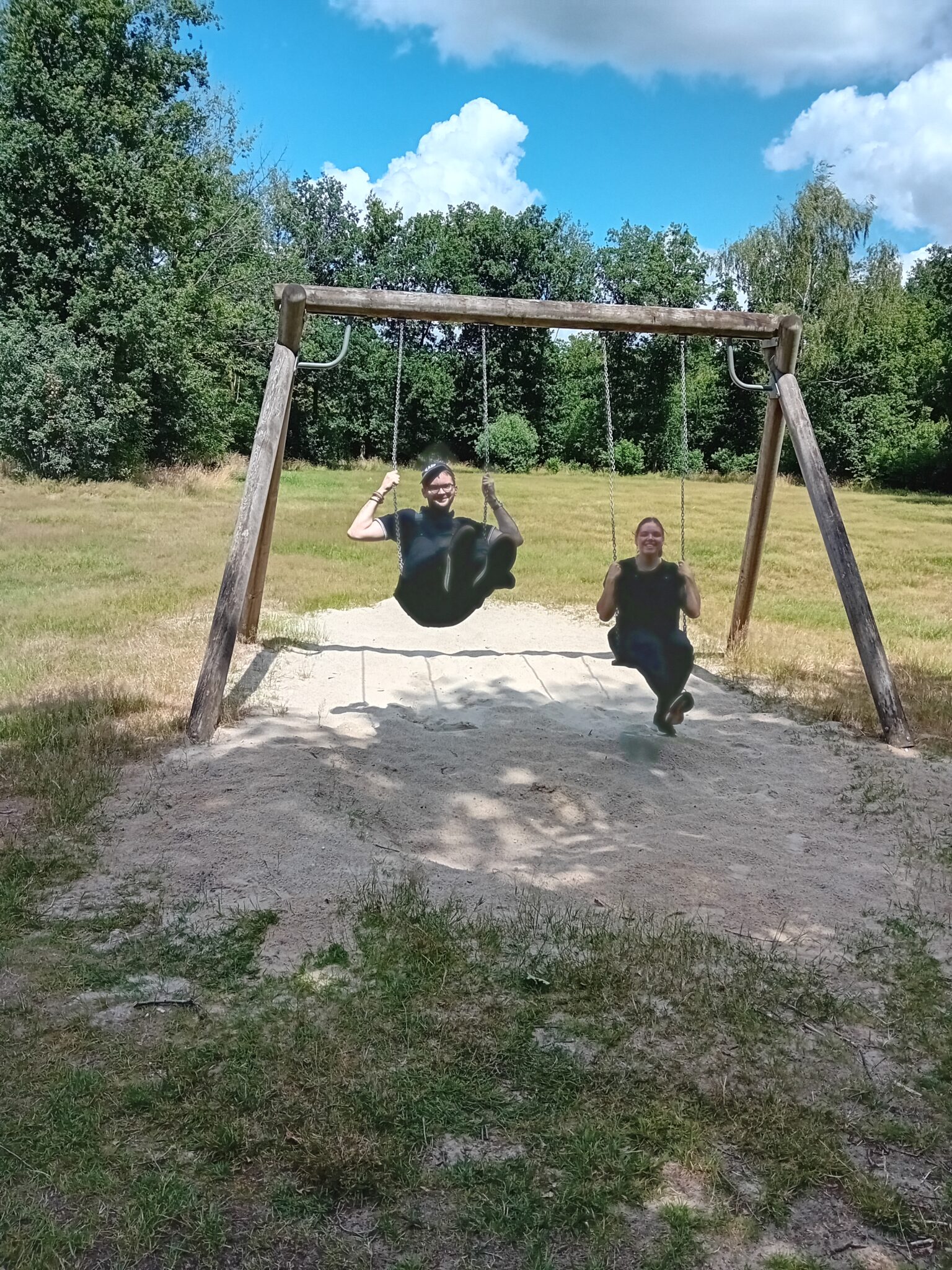 Twee mensen schommelen in een speeltuin omgeven door gras en bomen onder een blauwe hemel.
