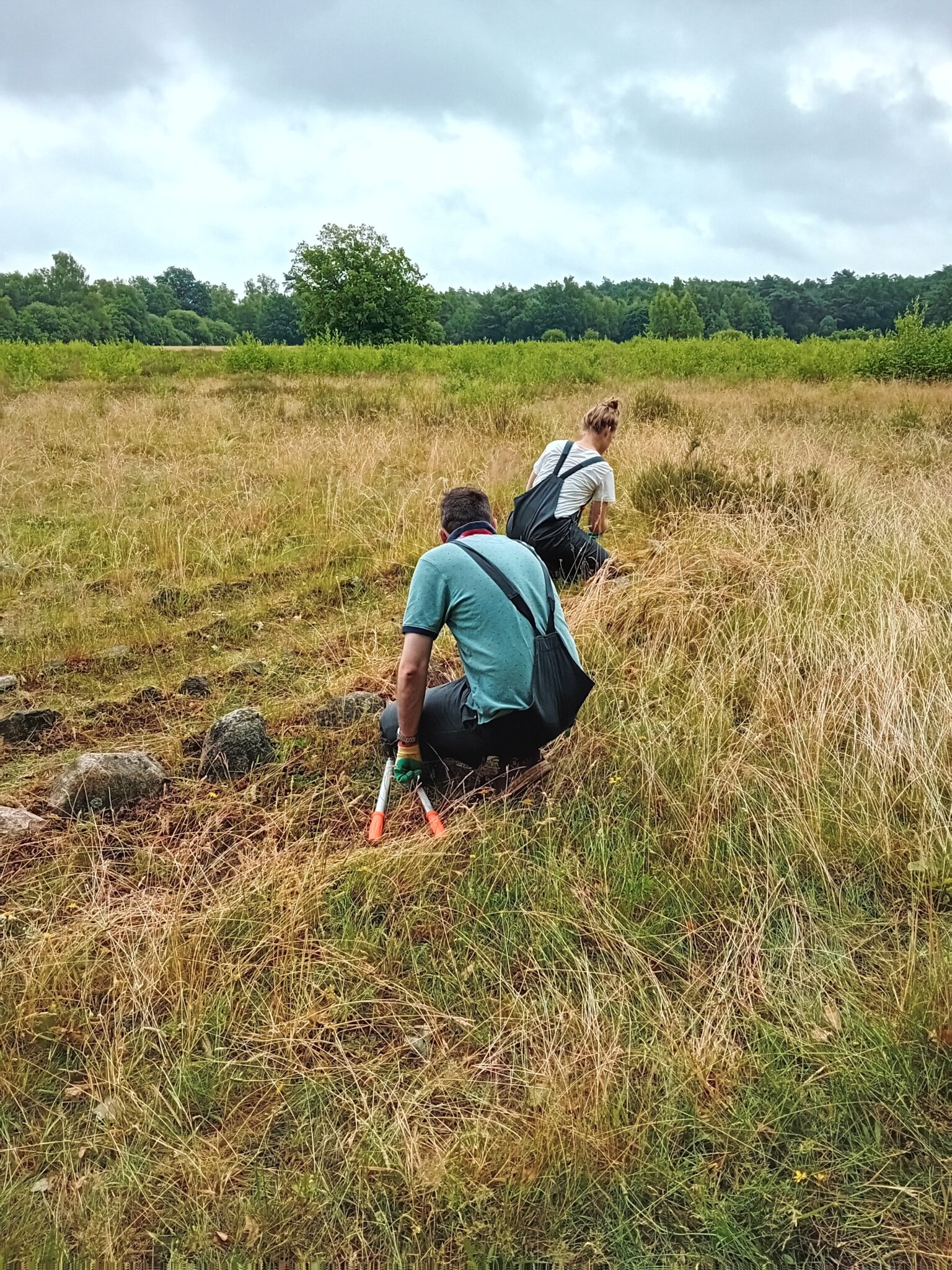 Twee mensen werken in een grasveld met gereedschap, omringd door natuur.