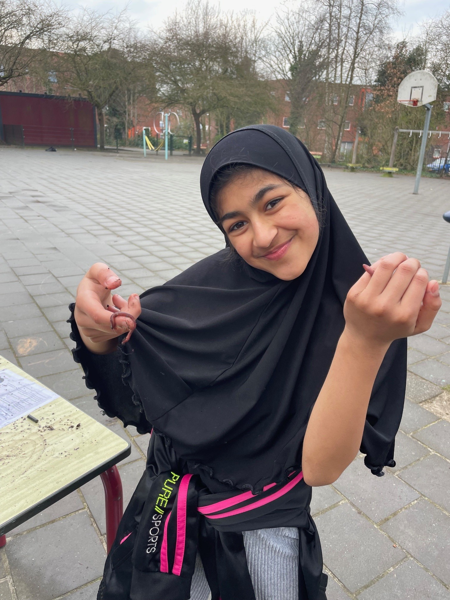 Meisje op schoolplein houdt lachend een worm vast, met een basketbalbord op de achtergrond.