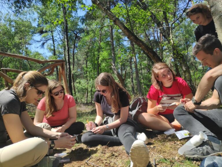 Groep jonge mensen zit samen in een bos, bezig met een activiteit op de grond.