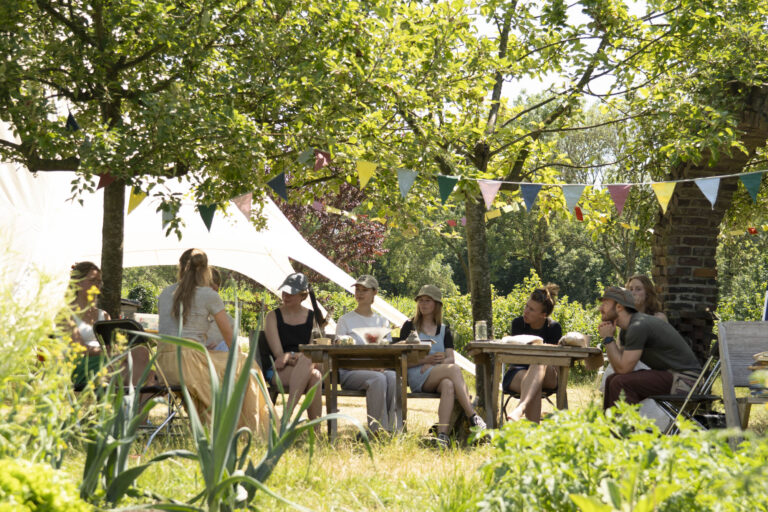 Groep mensen aan tafels in een zonnige tuin met vlaggetjes en schaduw van bomen.