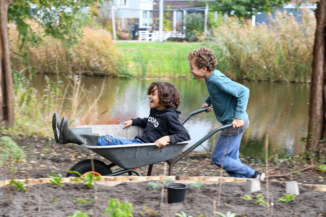 Twee kinderen spelen bij een waterkant, één rijdt in een kruiwagen, de ander duwt lachend.