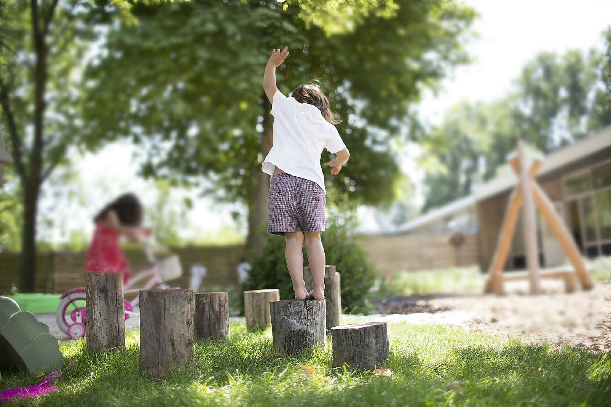 Kind speelt op houten blokken in een zonnige tuin, met ander kind op een fiets op de achtergrond.