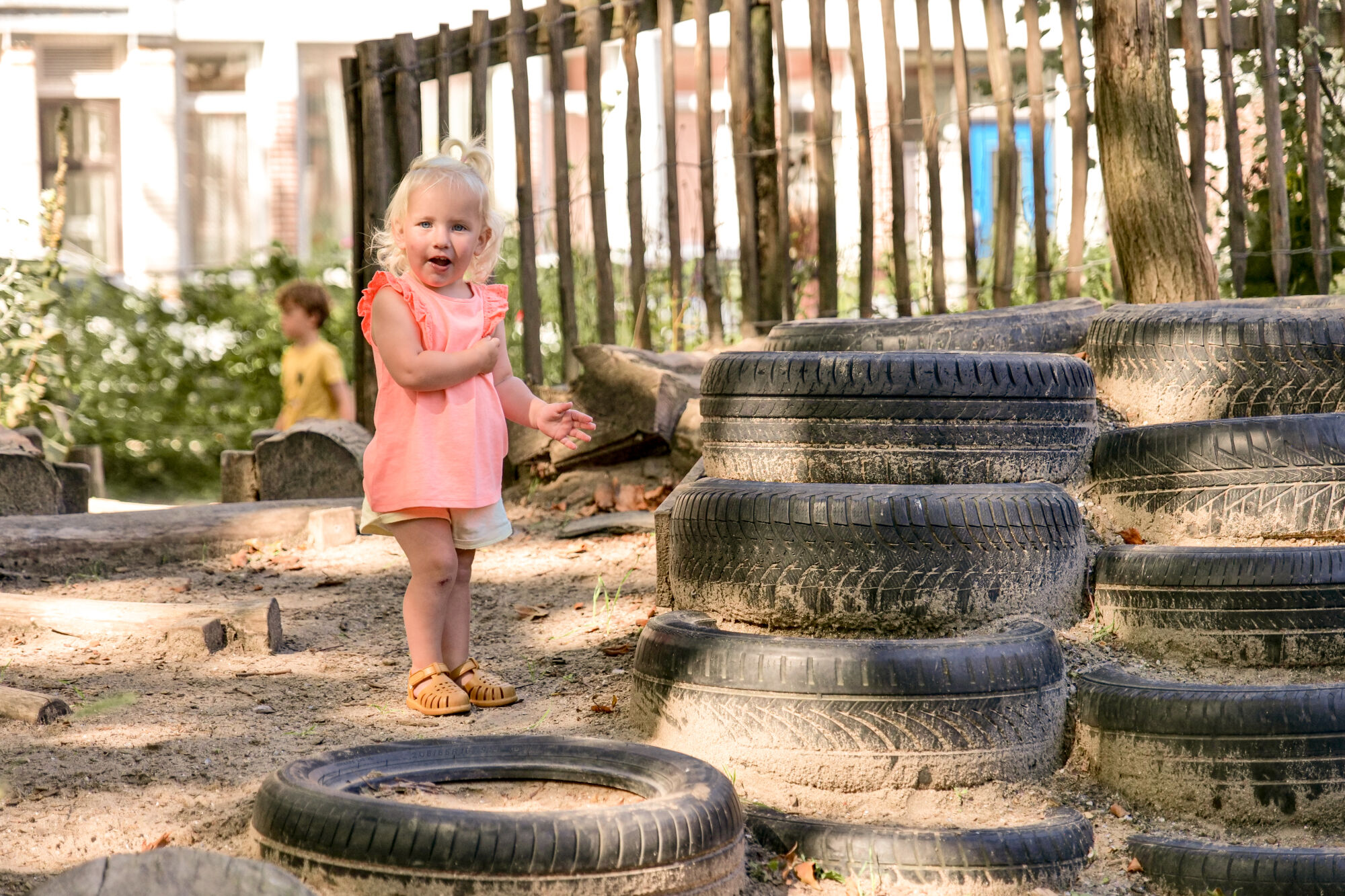 Peuter speelt bij een stapel autobanden in een zonnige, omheinde speeltuin.