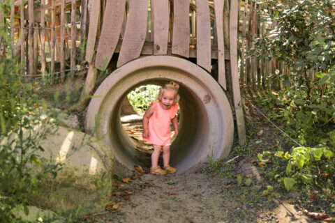 Kind in een roze jurk kruipt door een betonnen tunnel op een zonnige dag in een speeltuin.