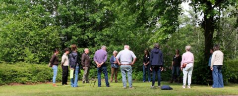 Groep mensen staat in een kring in een groene tuin, omringd door bomen en struiken.
