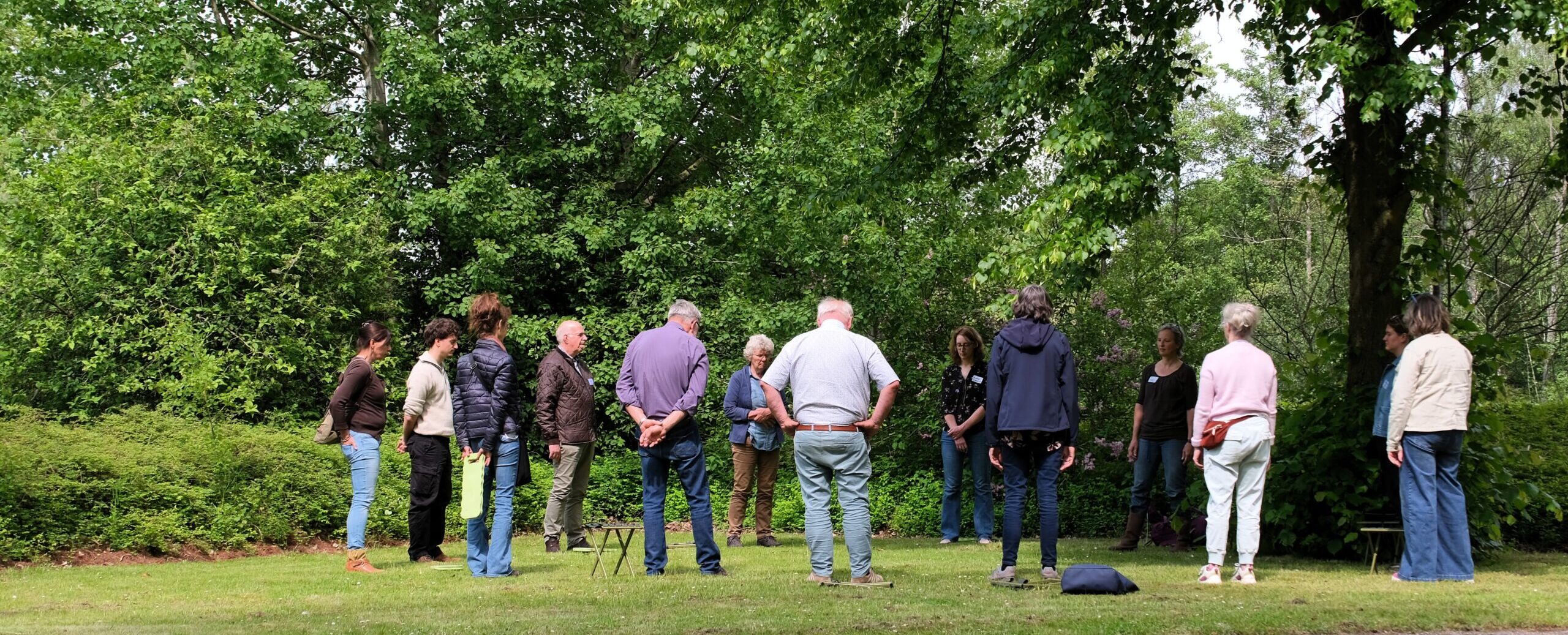 Groep mensen staat in een kring in een groene tuin, omringd door bomen en struiken.