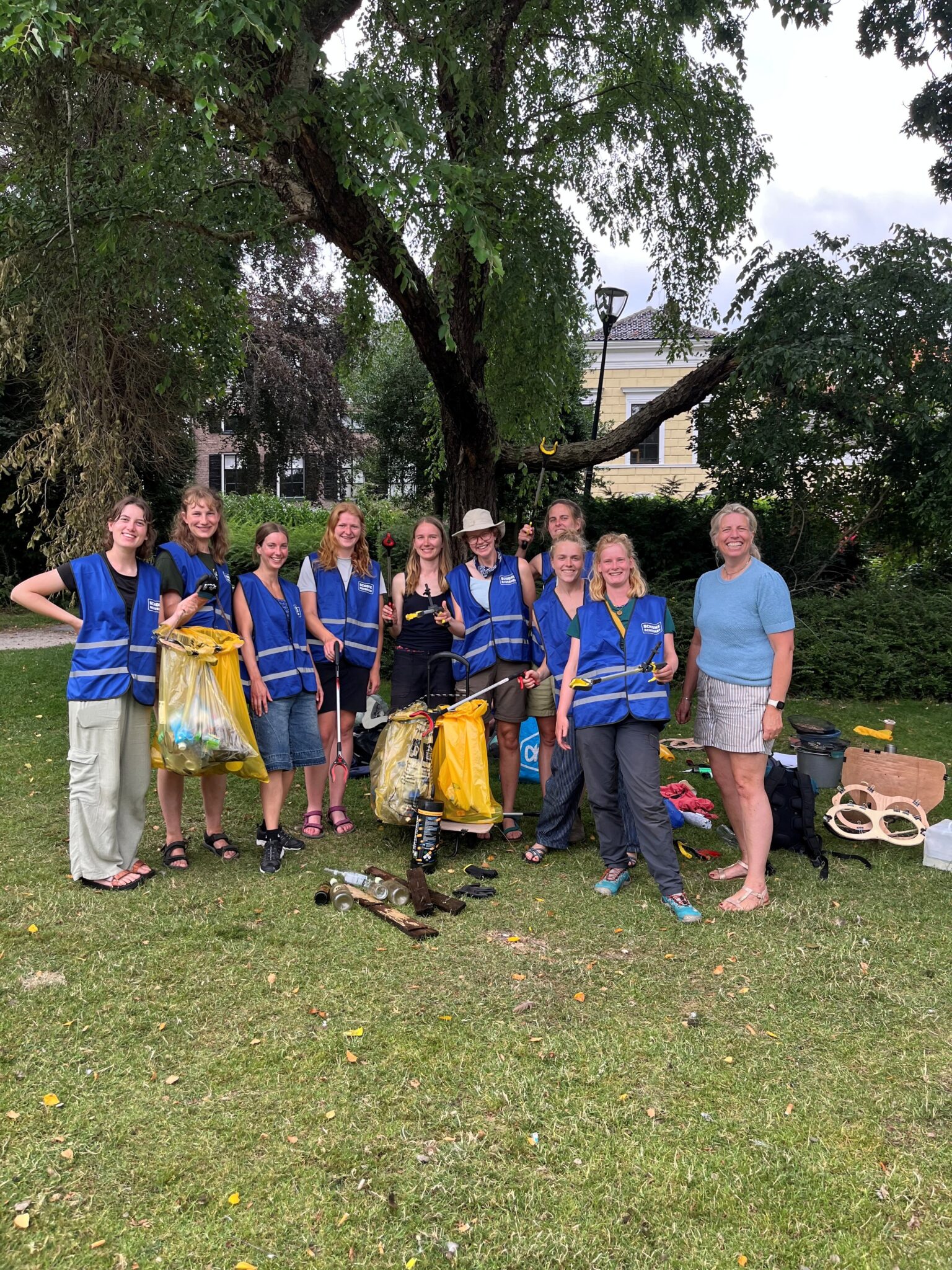 Groep mensen in blauwe hesjes poseert met vuilniszakken in een park na opruimactie.
