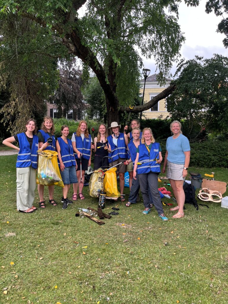 Groep mensen in blauwe hesjes poseert met vuilniszakken in een park na opruimactie.
