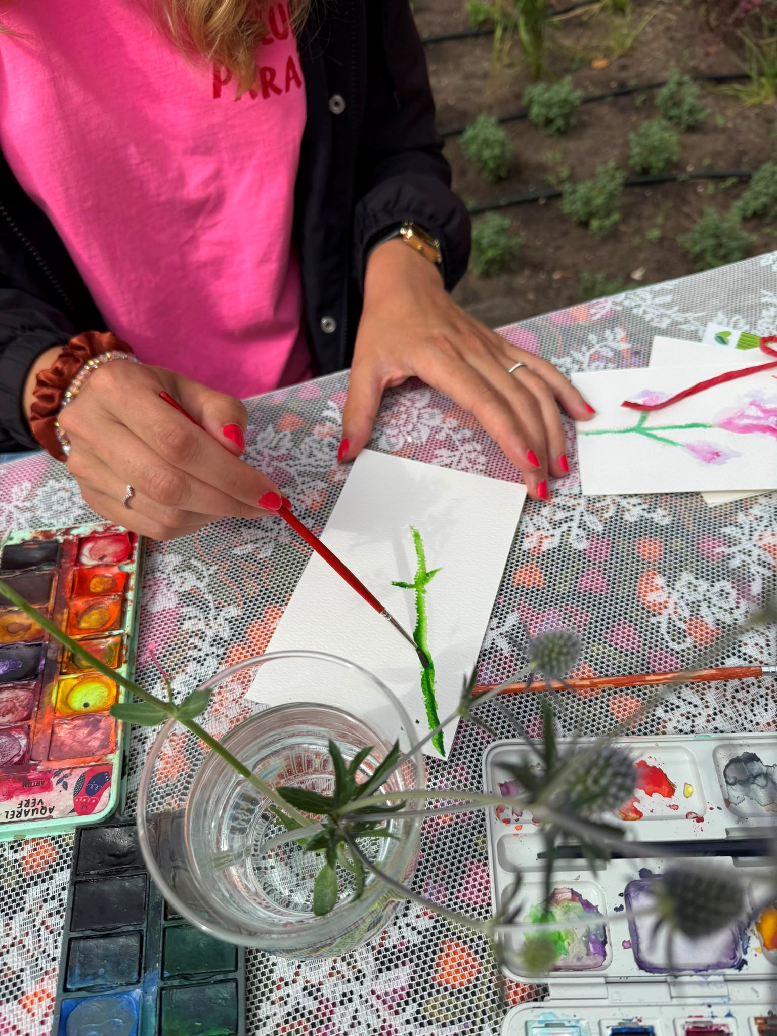 Persoon schildert bloemen met waterverf op papier, bloemen in glas, kleurrijke pallet op tafel.