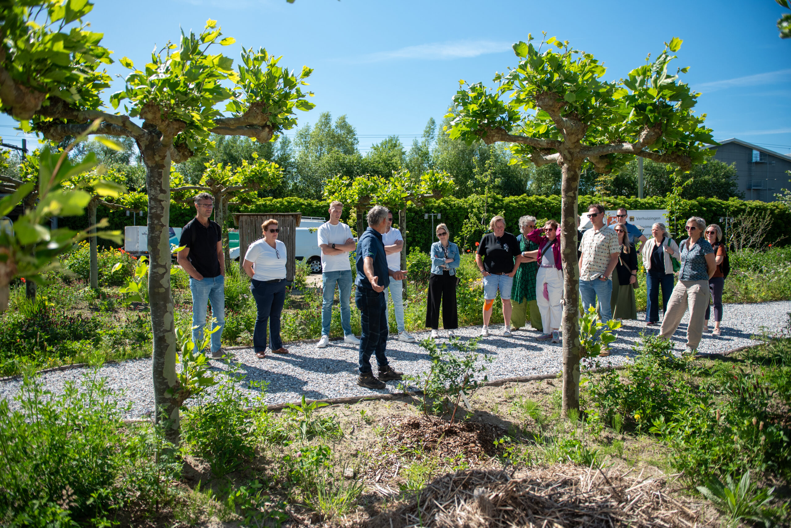 Groep mensen luistert naar een gids in een zonnige, groene tuin met bomen.