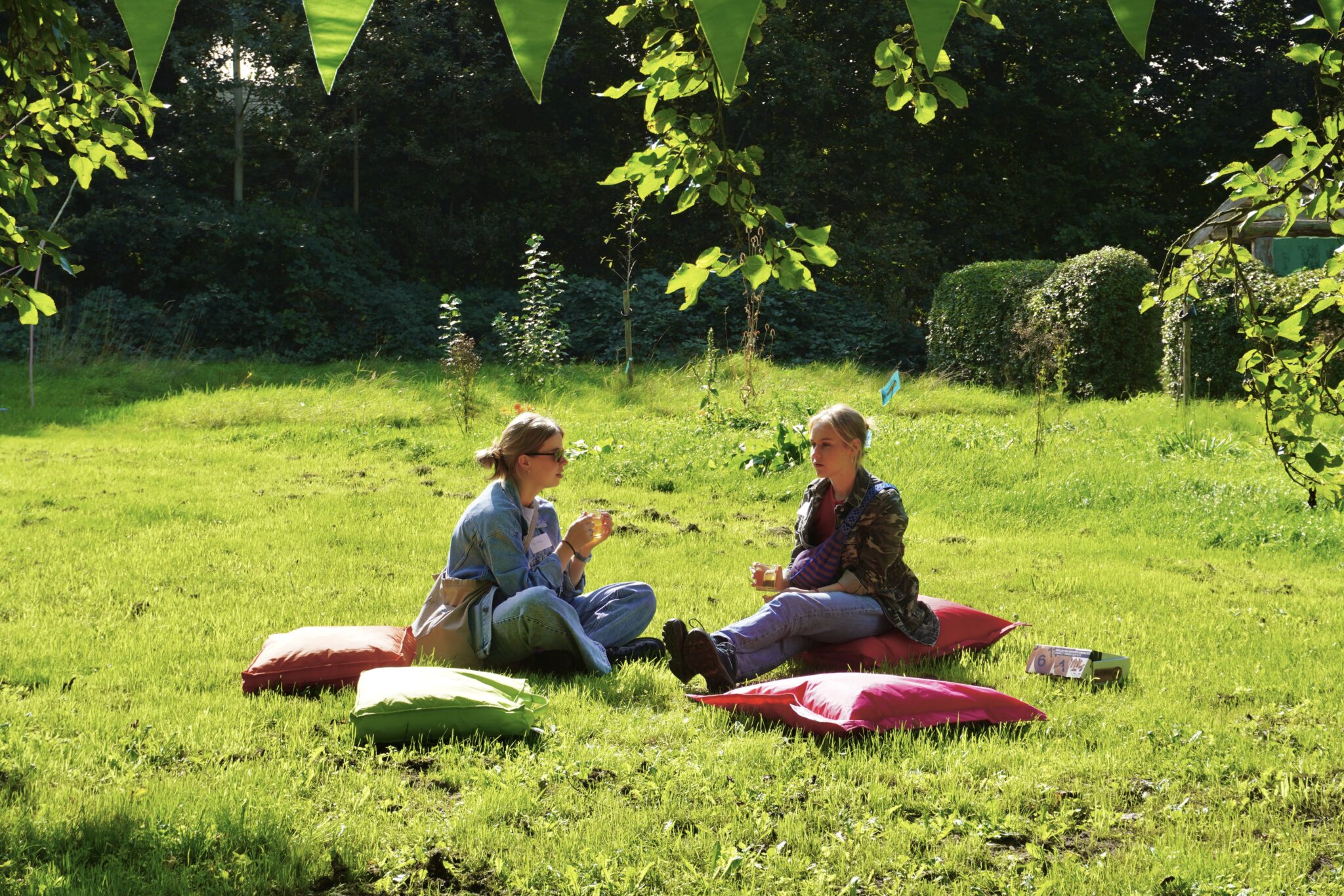 Twee mensen zitten op kussens in een zonnige tuin, omgeven door groen, met kopjes in hun hand.