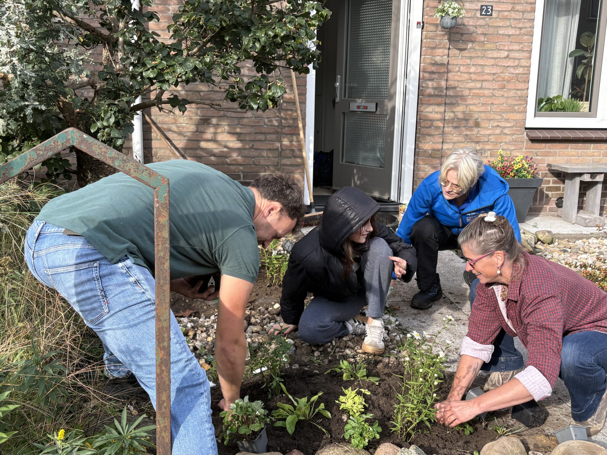 Mensen planten samen bloemen voor een huis met bakstenen muur en groene planten.