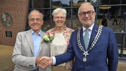 Twee mannen schudden handen, vrouw tussen hen in. Man rechts draagt ketting, mogelijk een burgemeester.