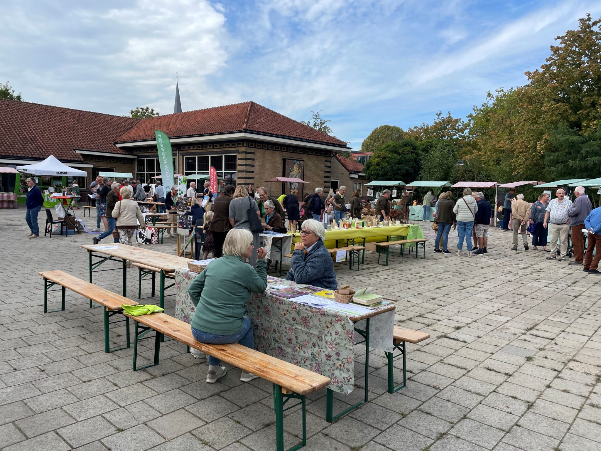 Buitenkraampjesmarkt met mensen en tafels op een plein bij een gebouw, omgeven door bomen.