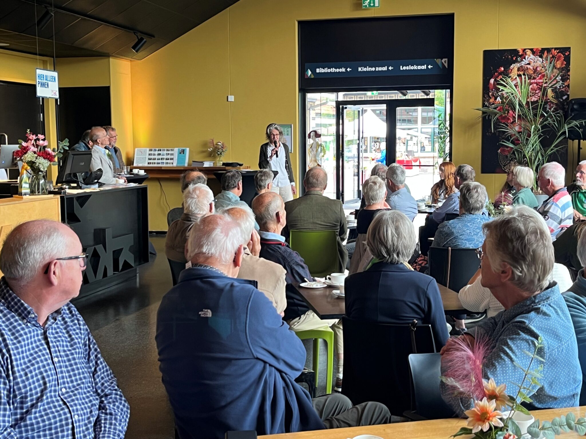 Groep ouderen luistert naar spreker in een zaal, met bloemen en een infobalie op de achtergrond.