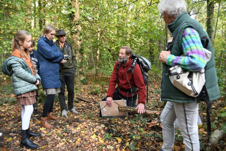Groep mensen in een bos, luisterend naar een knielende gids met rugzak.