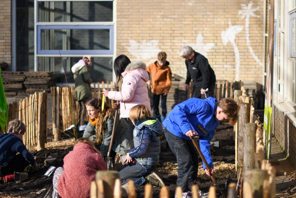 Mensen planten samen bomen in een afgebakend stuk aarde bij een gebouw met een muurschildering.