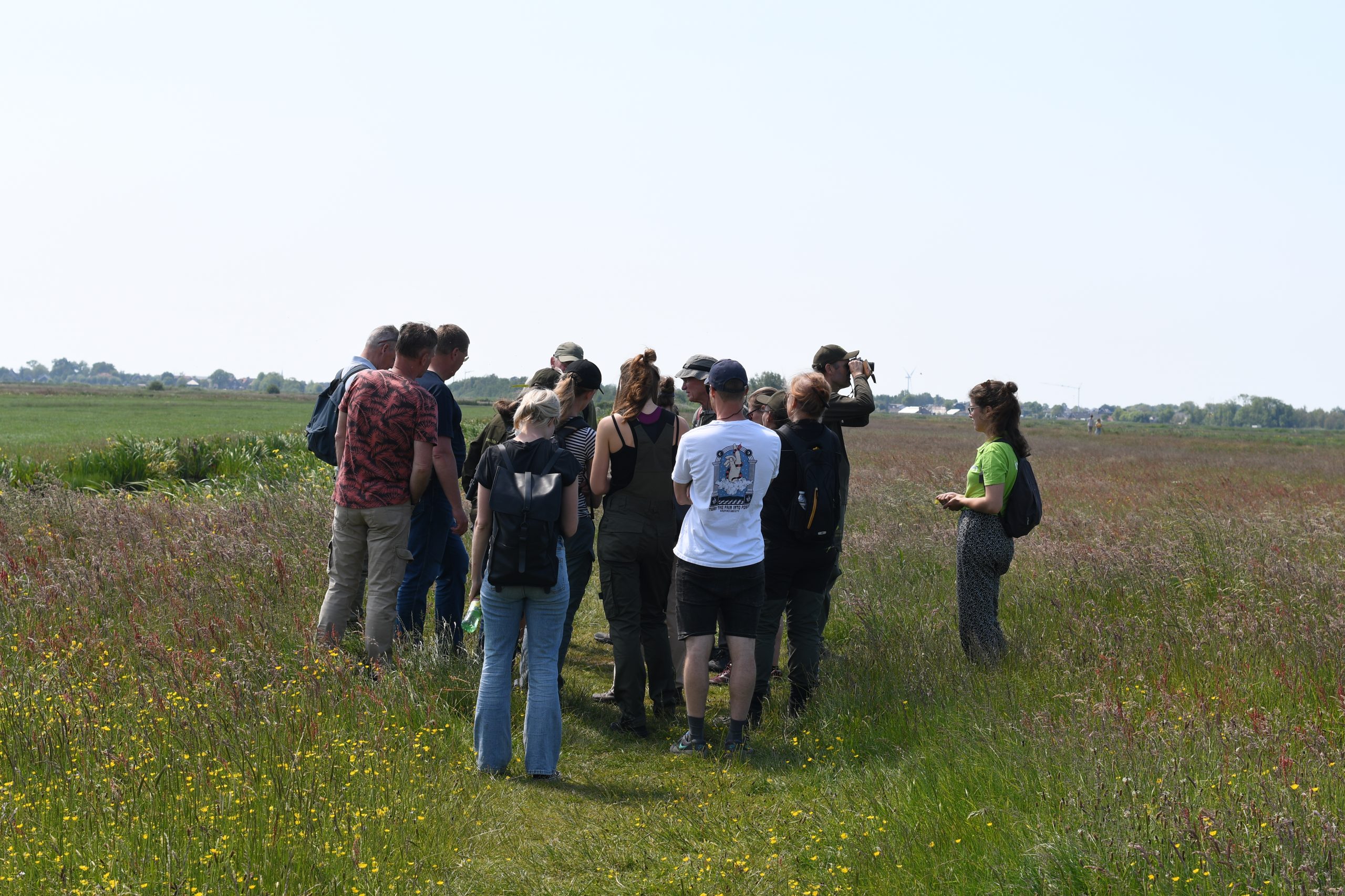 Groep mensen met rugzakken wandelt door een bloemrijk grasland onder een heldere lucht.