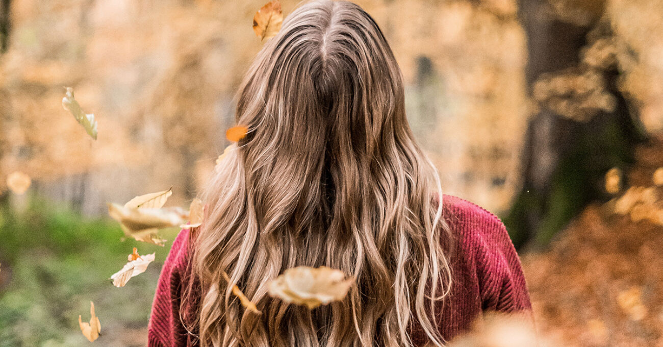 Een vrouw met lang haar loopt in een herfstbos, omgeven door vallende bladeren.