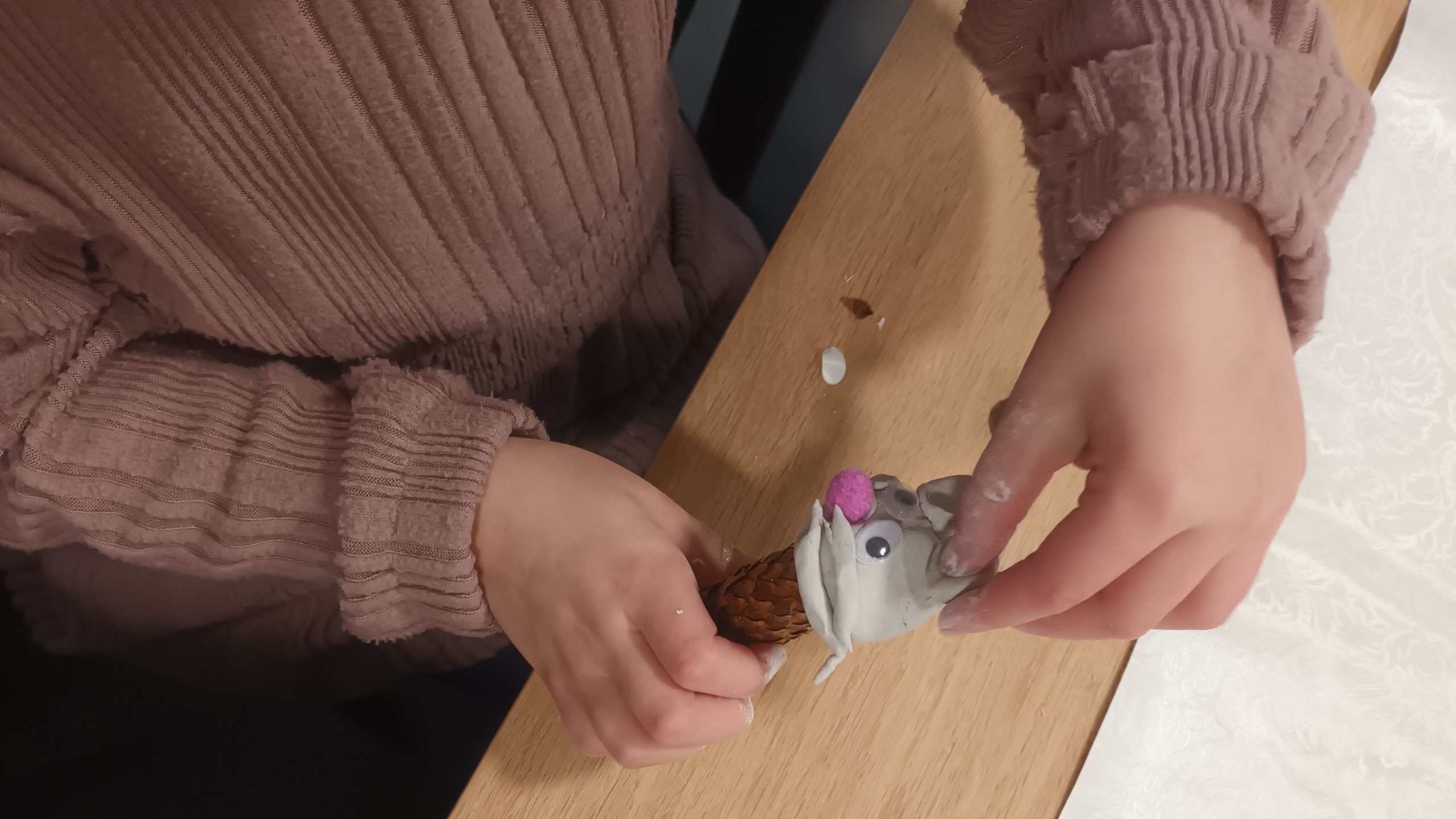 Een kind knutselt met een dennenappel, klei en googly eyes aan een houten tafel.