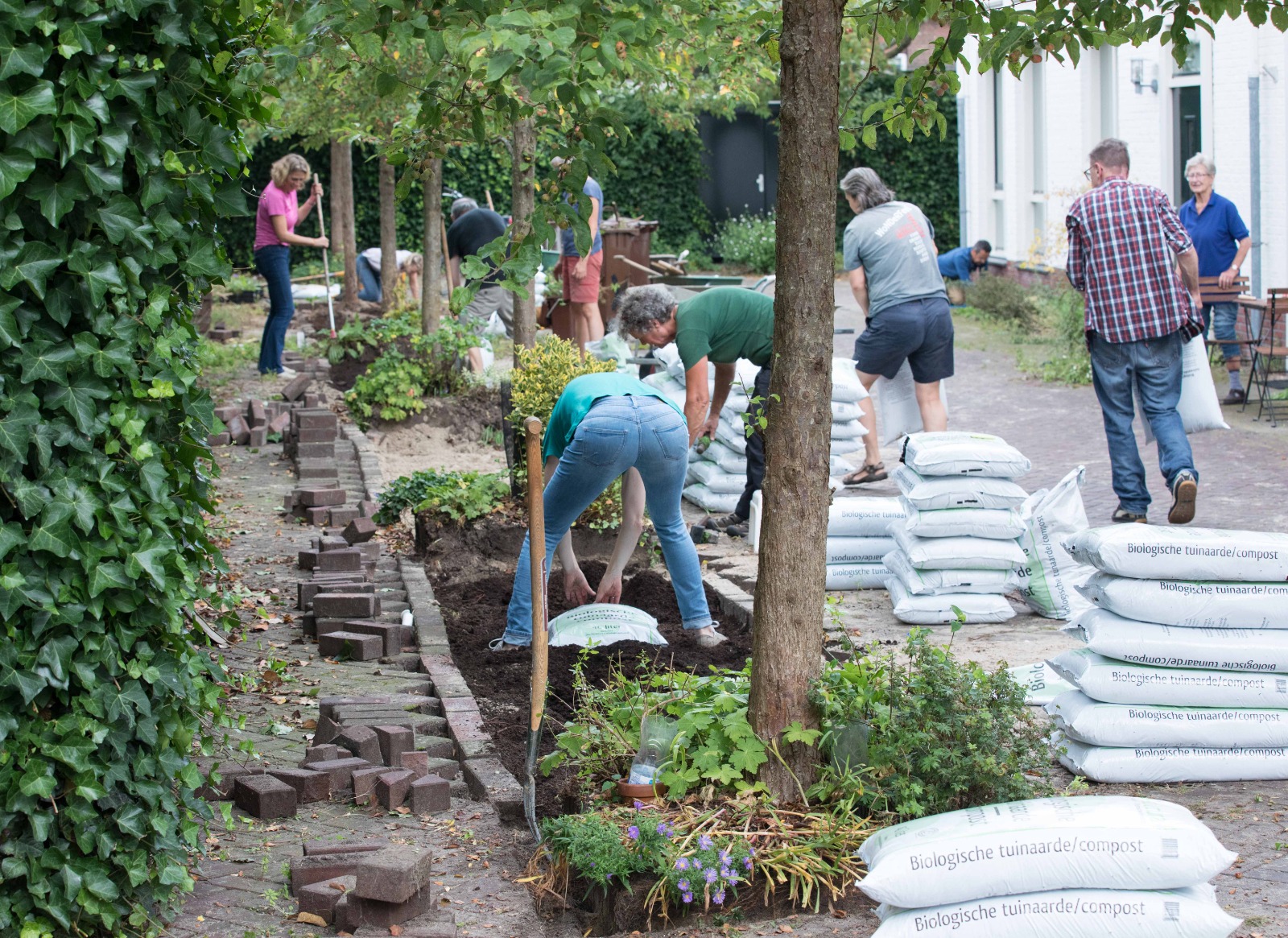 Mensen werken in een tuin met stapels biologische compostzakken omringd door bomen.