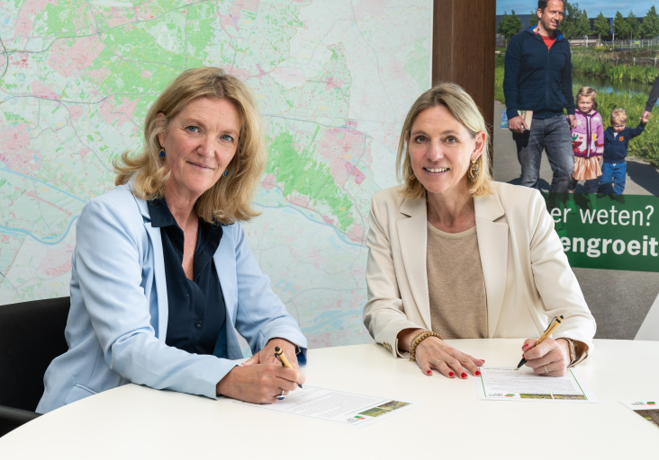 Twee vrouwen ondertekenen documenten aan een tafel; achtergrond met een kaart en een poster.