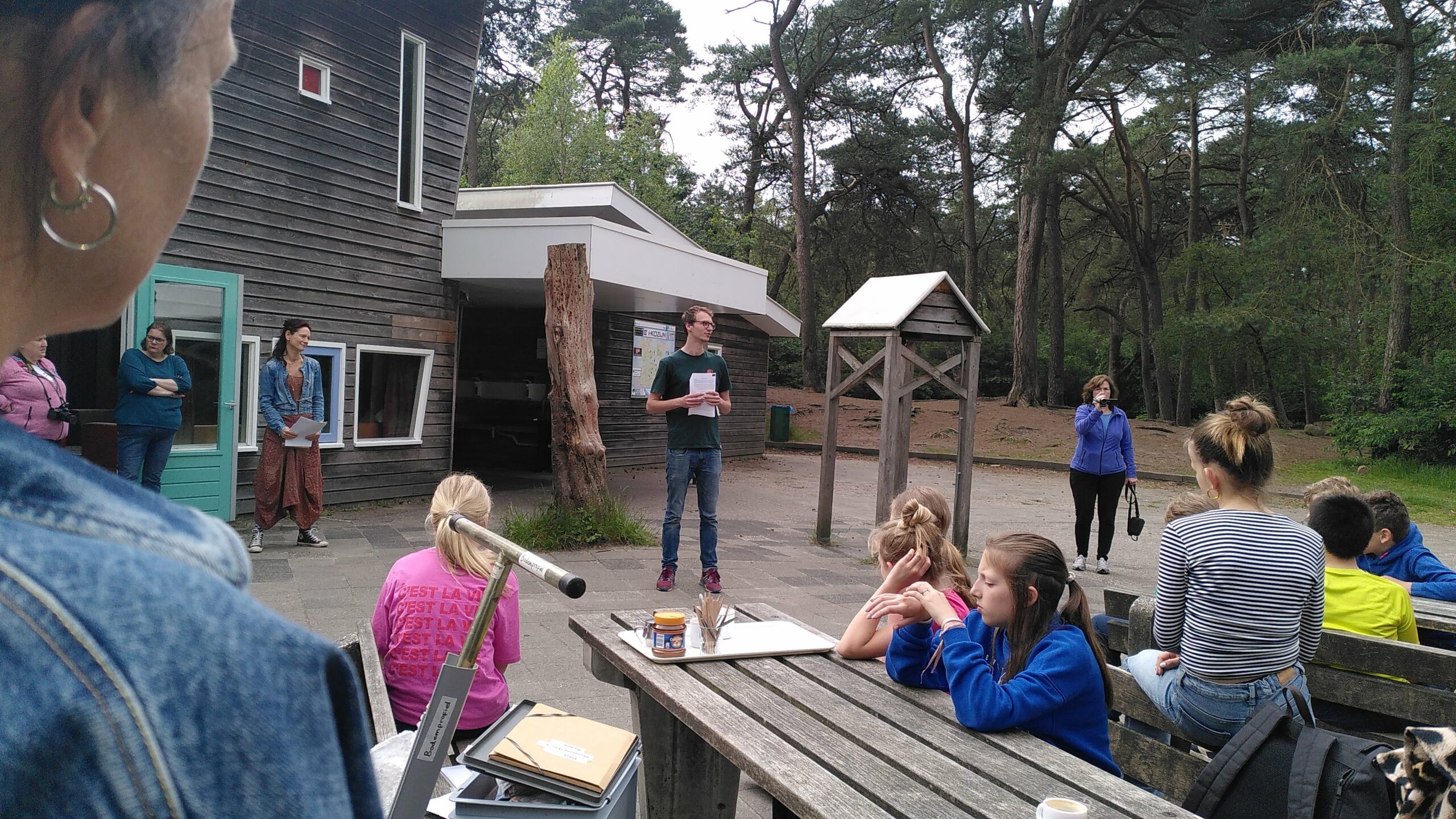 Groep mensen luistert naar spreker buiten bij houten gebouw, omringd door bomen, met picknickbank.