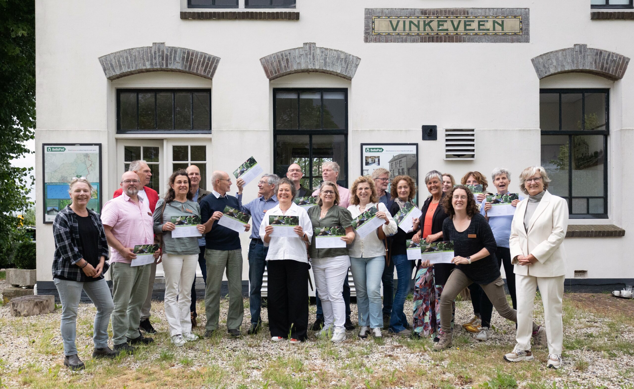 Groep mensen poseert met certificaten voor een gebouw met het bord "Vinkeveen".