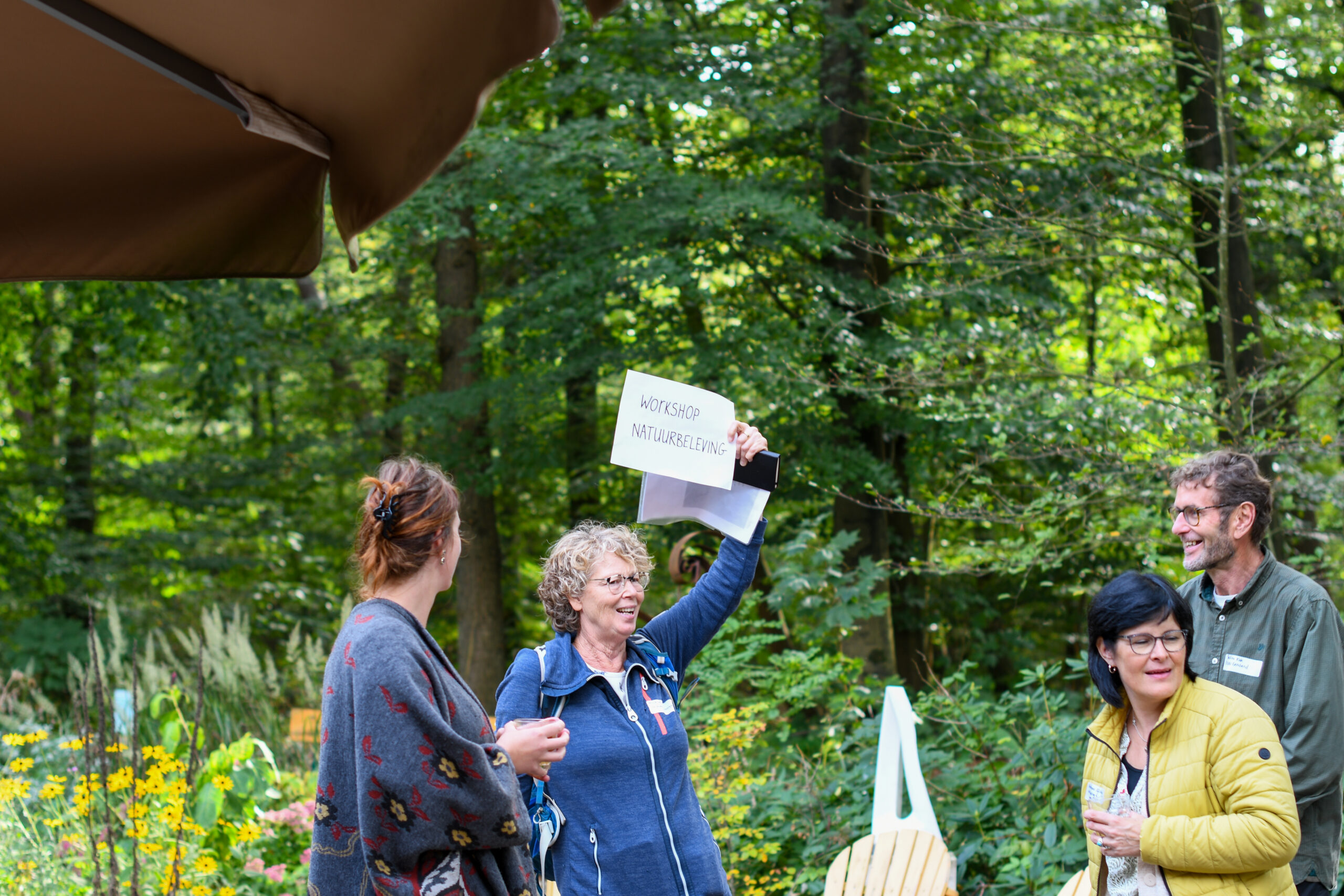 Vier mensen in een bos, vrouw houdt bord "Workshop Natuurbeleving" omhoog.