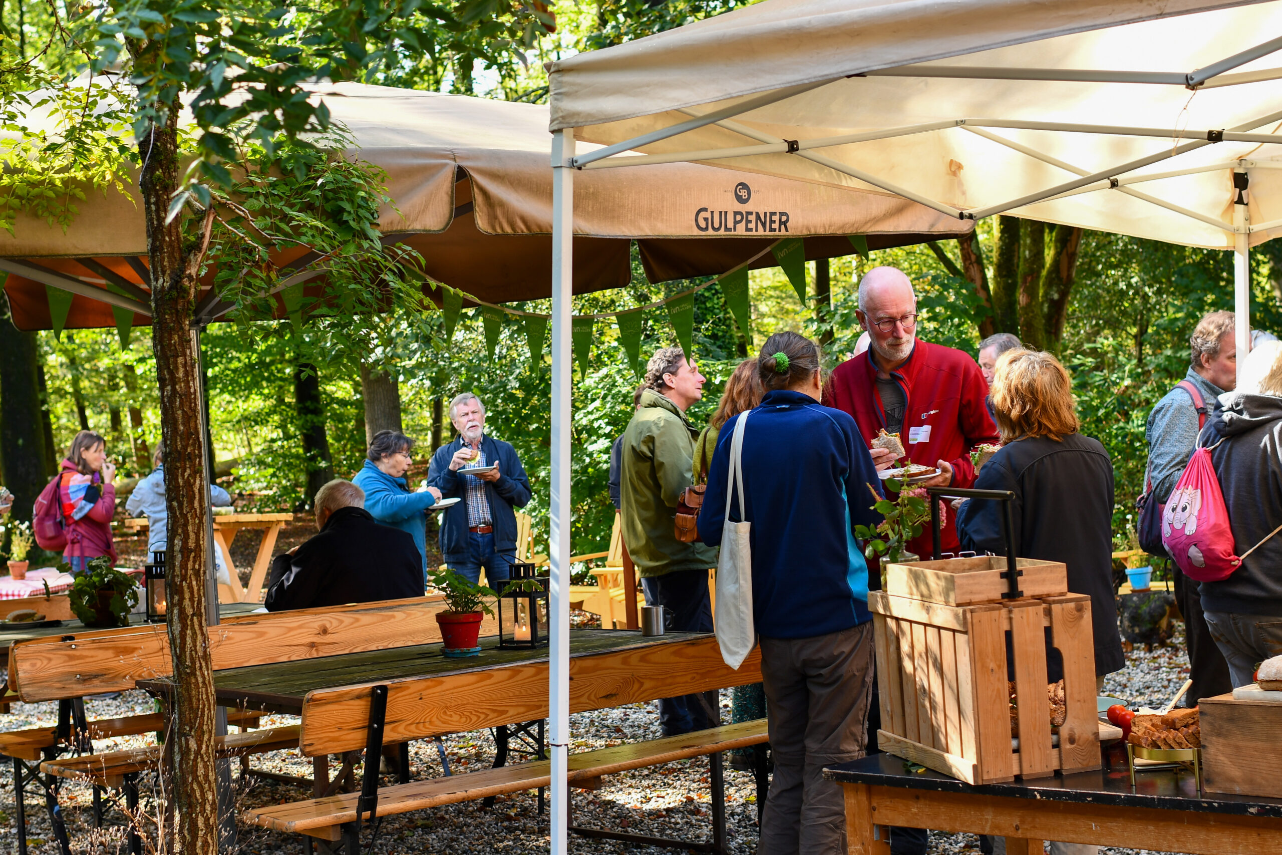 Groep mensen onder tenten in een groene buitenomgeving, genietend van eten en drinken.