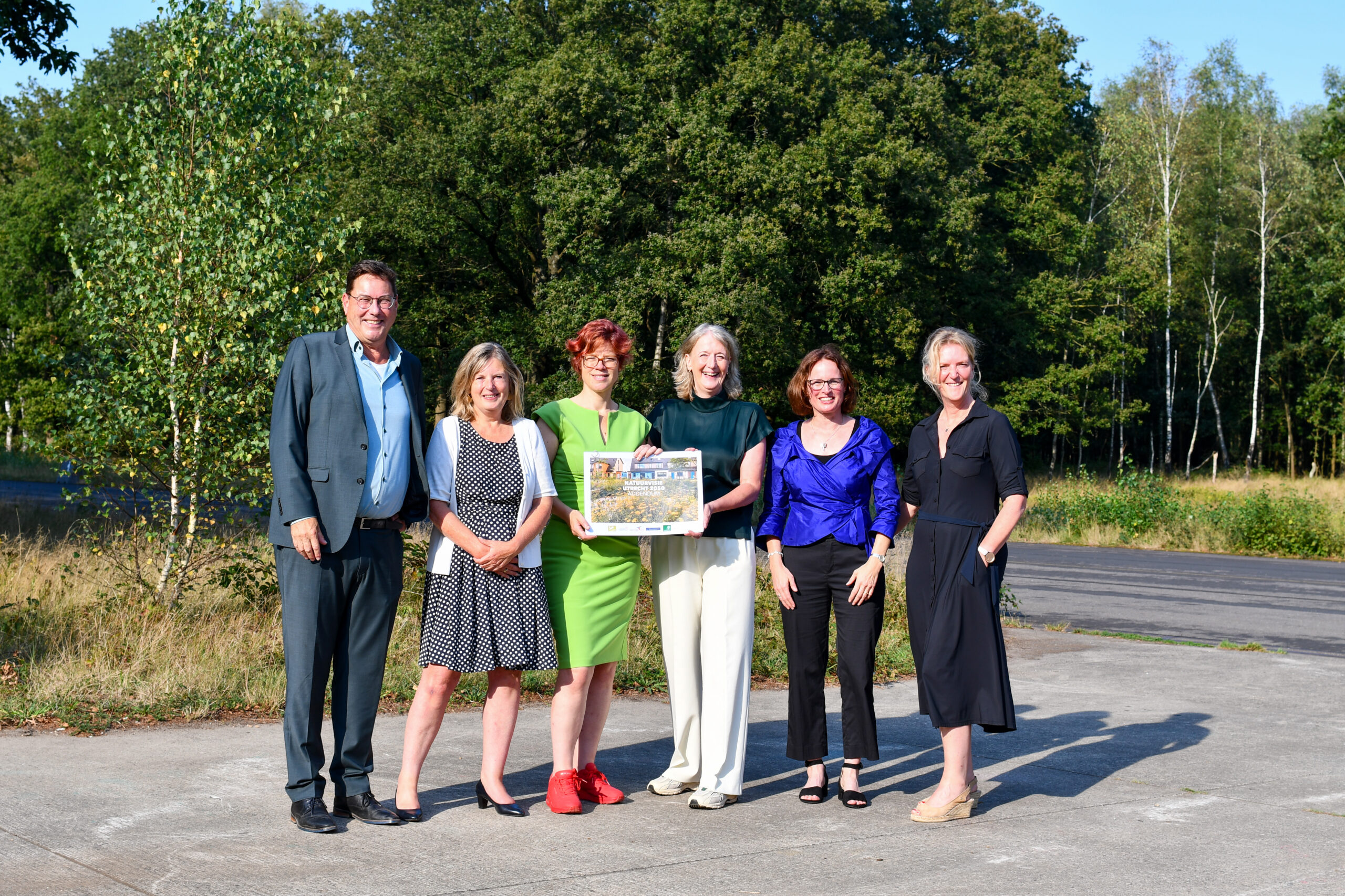 Zes mensen poseren buiten met een papier, tegen een achtergrond van bomen en gras.