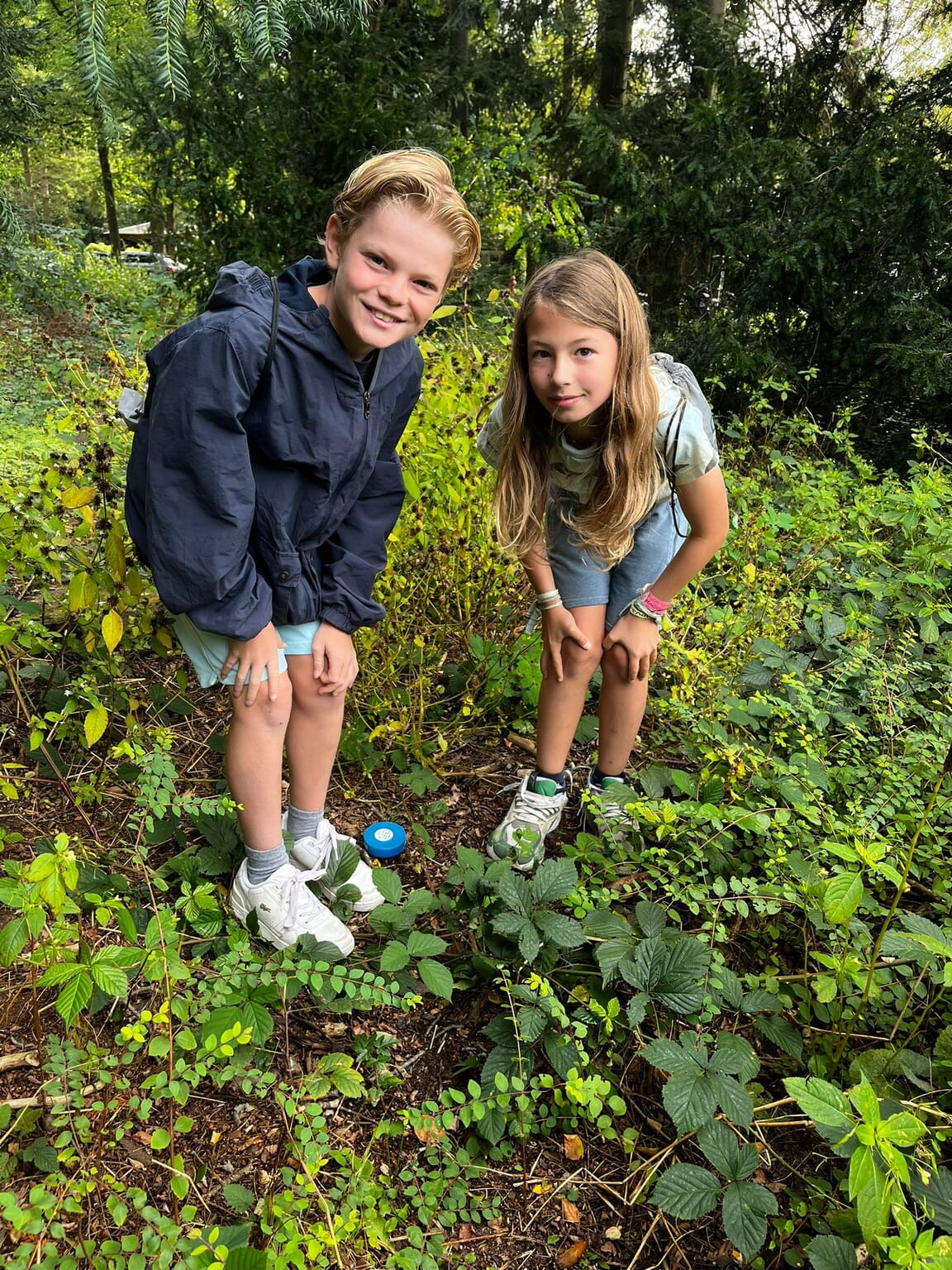 Twee kinderen staan buiten in een bosrijke omgeving, gebogen over een blauw bakje op de grond.