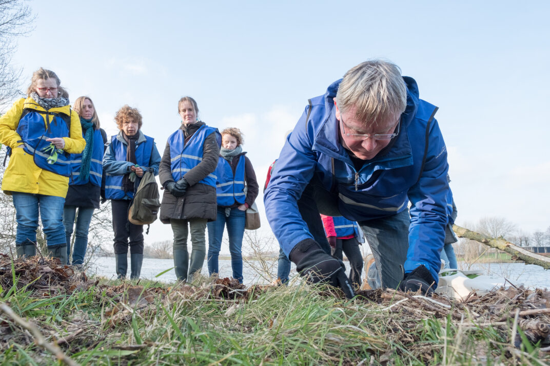 Mensen in blauwe hesjes observeren een man die op de grond naar iets zoekt in een natuurlijke omgeving.