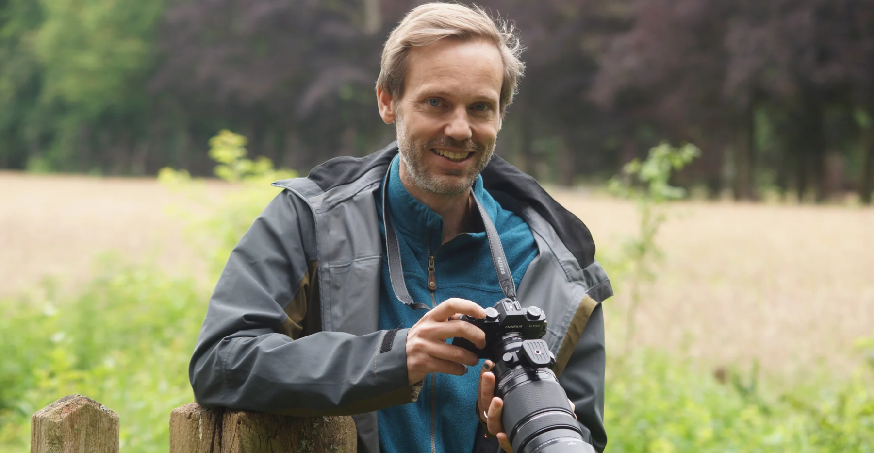 Man in een jas met een camera, glimlachend in een groene, bosrijke omgeving.