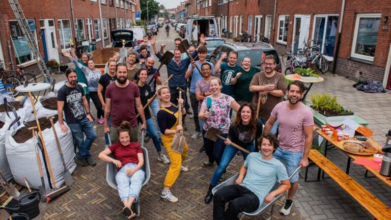 Groepsfoto van blije mensen in een straat tijdens een buurtproject, met gereedschap en planten.