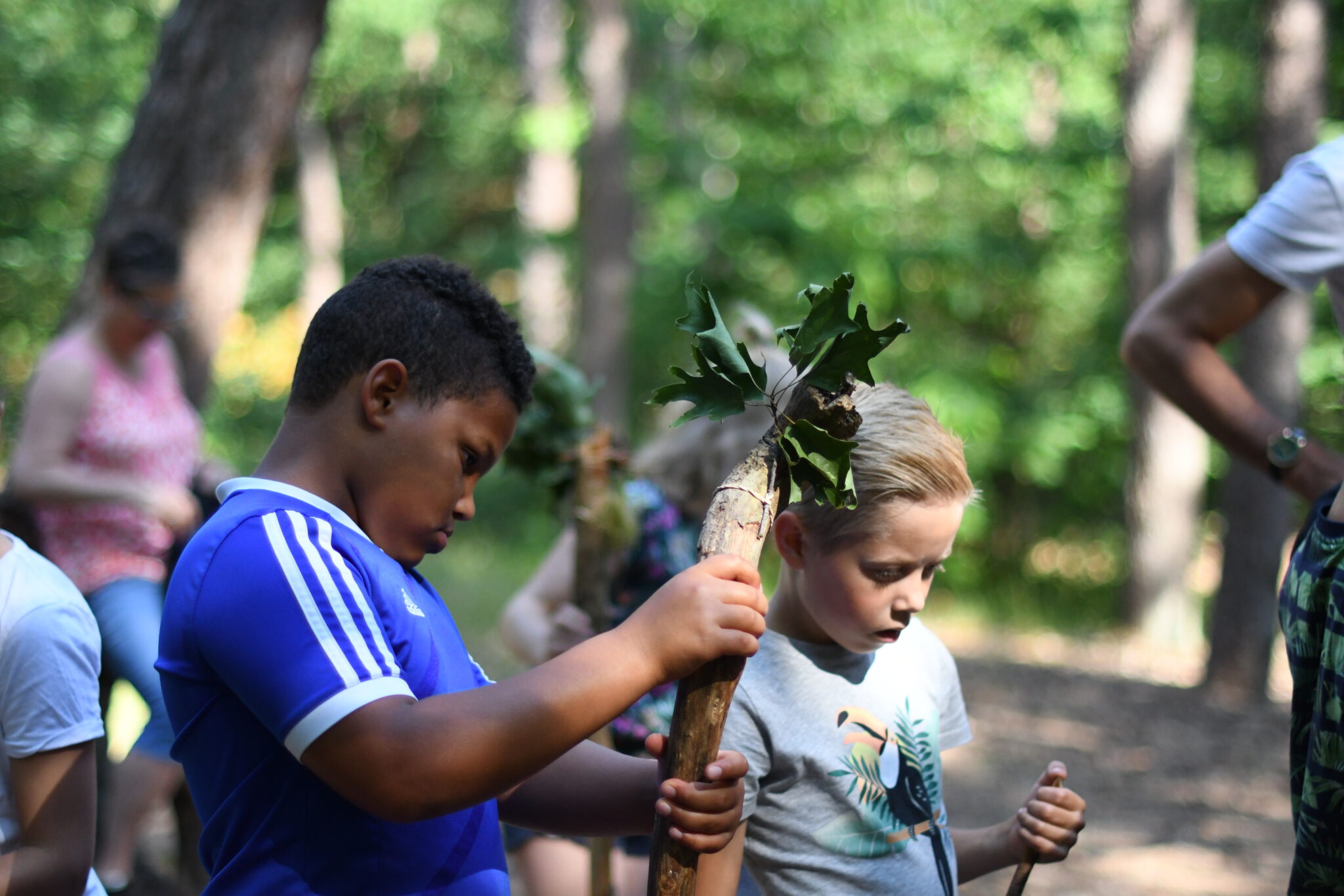 Twee kinderen met takken en bladeren in een bosrijke omgeving.