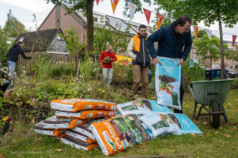 Mensen werken in een tuin met zakken compost en een kruiwagen bij een huis met vlaggetjes erboven.