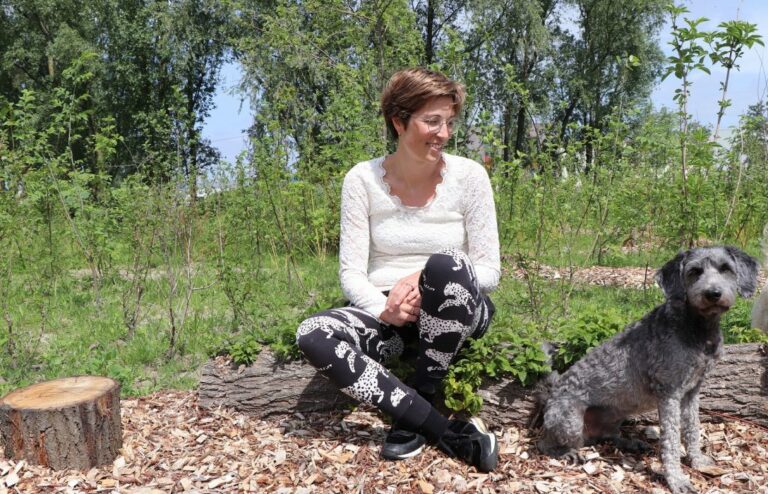 Vrouw zit op een boomstam naast een grijze hond, omringd door groen in een parkachtige omgeving.