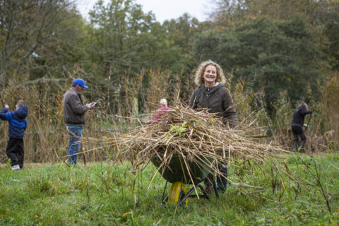 Persoon met kruiwagen vol takken, andere mensen werken of kijken op hun telefoon in een groen natuurgebied.