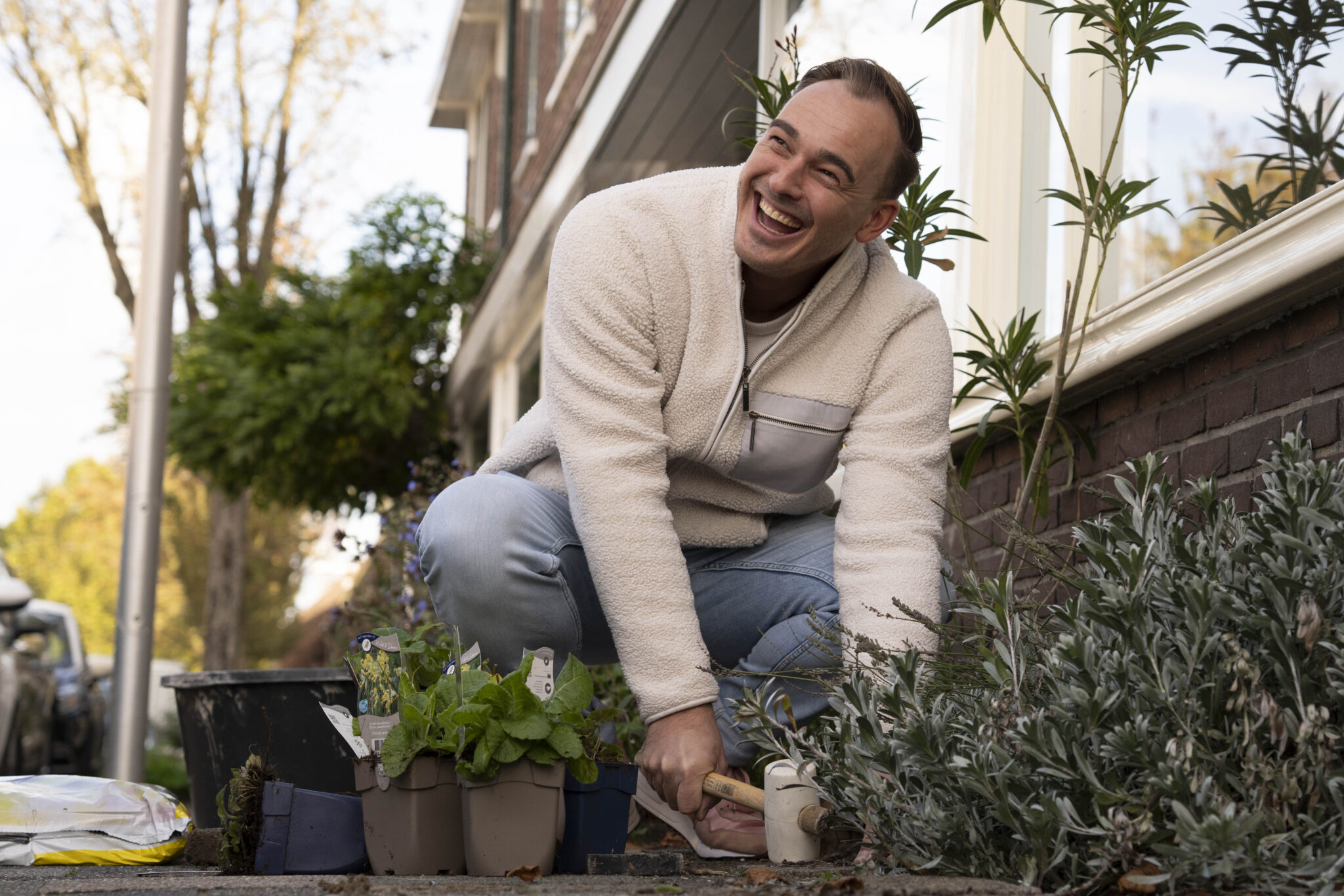 Man in witte trui plant bloemen buiten en lacht vrolijk.