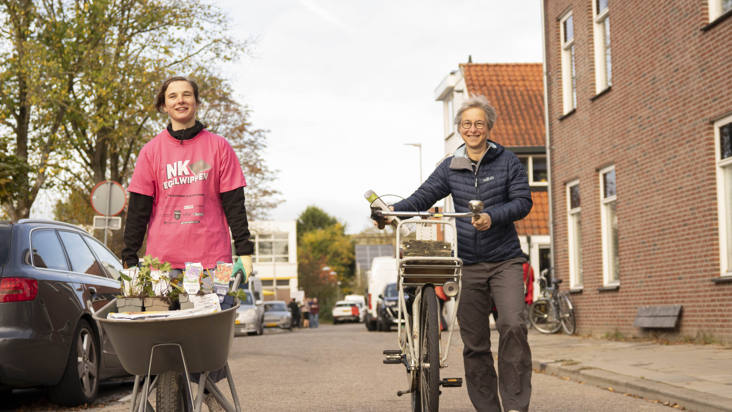 Twee mensen in een straat, één met een kruiwagen vol planten en een ander met een fiets.