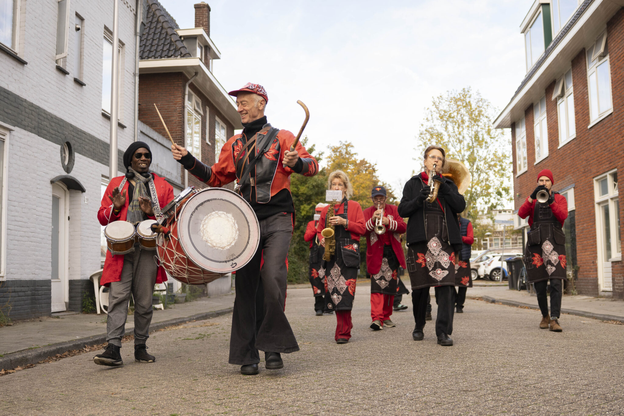 Straatfanfare in rode kleding, met drums en blaasinstrumenten, marcheert door een woonwijk.