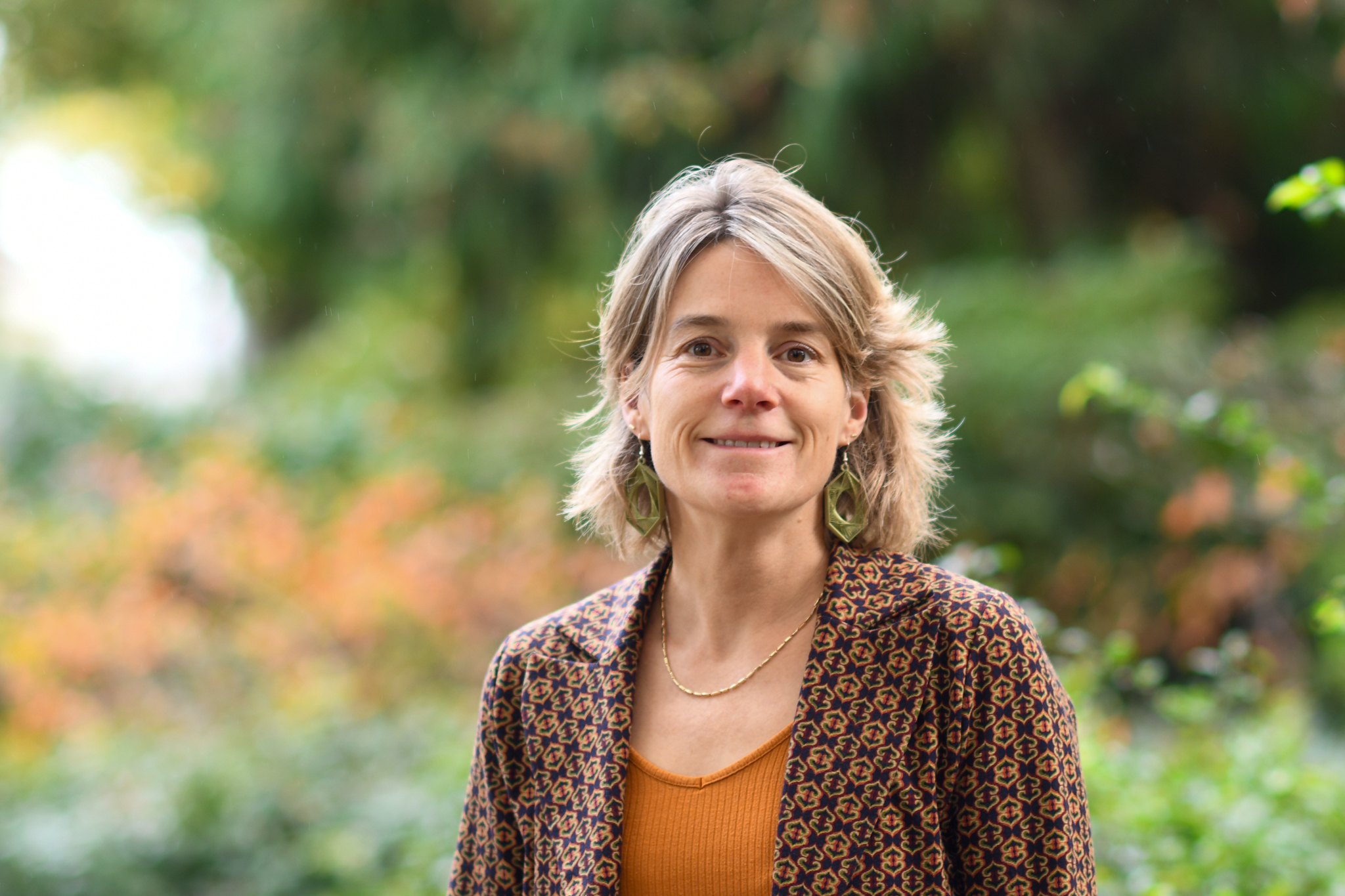 Vrouw glimlacht buiten, omringd door groene en herfstige planten.