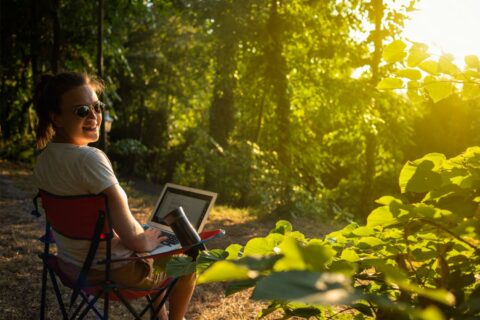 Vrouw met laptop in stoel in zonnig bos, glimlachend naar de camera.