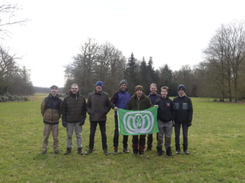 Groep van acht mensen op een grasveld met een groene vlag en bomen op de achtergrond.