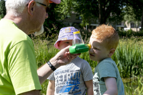 Opa laat kinderen een waterdiertje bekijken door een vergrootglas in een park.