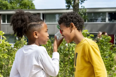 Twee kinderen onderzoeken een potje buiten, met groen en een gebouw op de achtergrond.