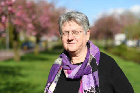 Vrouw met grijs haar, paarse sjaal, buiten met bloeiende bomen en blauwe lucht.