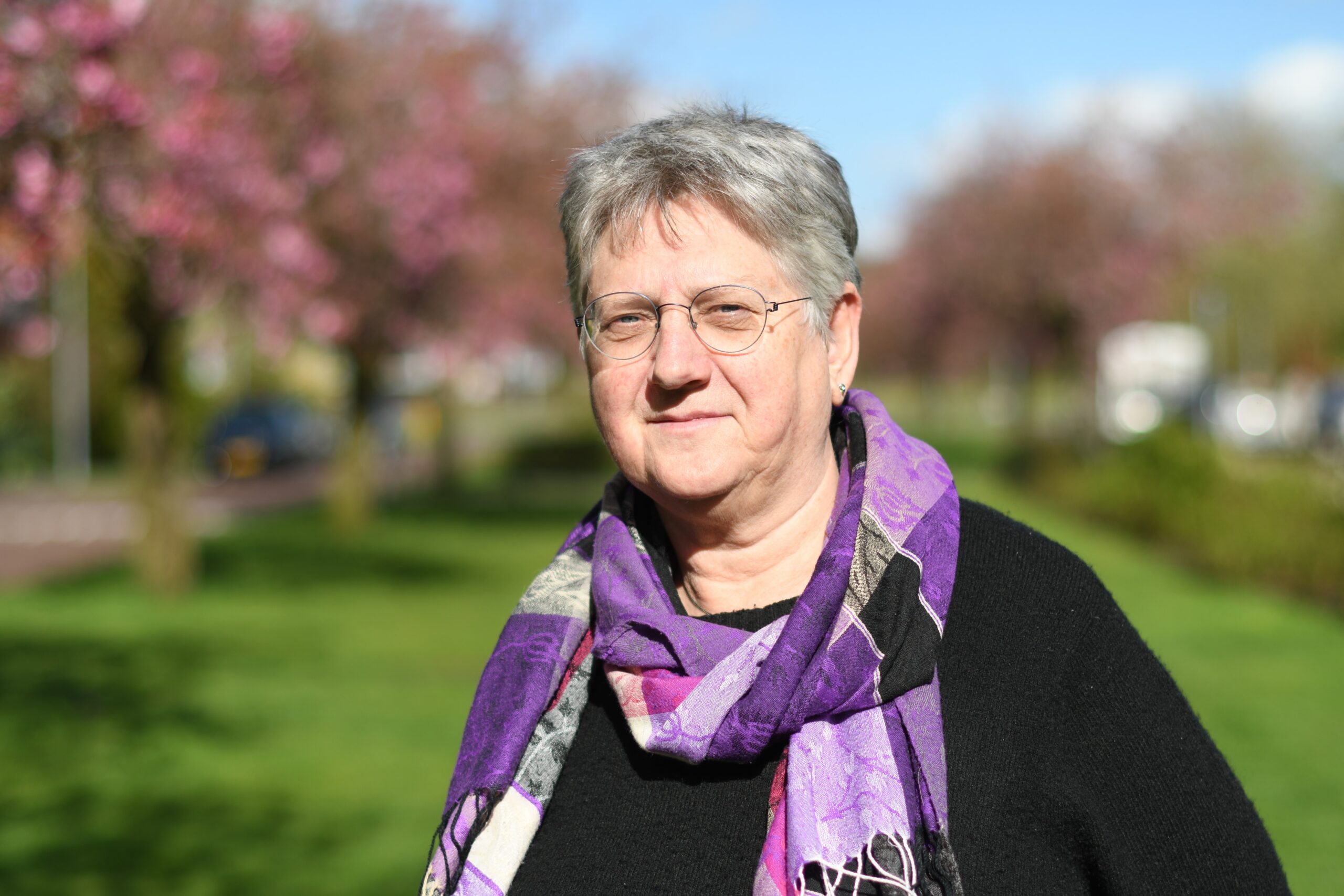 Vrouw met grijs haar, paarse sjaal, buiten met bloeiende bomen en blauwe lucht.